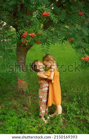 Girl sweetly and tenderly kisses her older sister on the forehead. Love and friendship between children. Tenderness in relationships. Kids in the garden by a tree with rowan berries