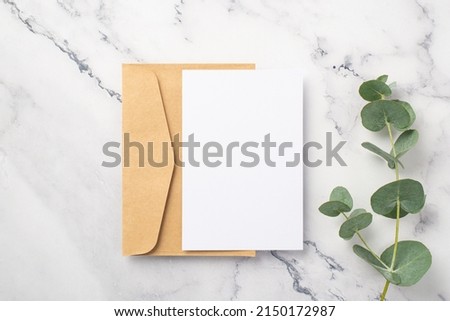 Top view photo of craft paper envelope paper card and eucalyptus sprig on white marble background with copyspace