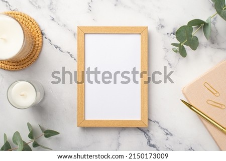 Top view photo of wooden photo frame candles on rattan serving mat pink diary gold pen clips and eucalyptus on white marble background with empty space