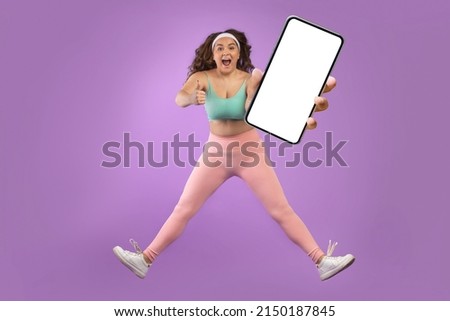 Satisfied excited young european plus size lady with open mouth in sportswear jump, freeze in air, show phone with blank screen, isolated on purple background. Blog, app, great result and health care