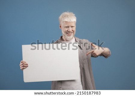 Place for your ad. Excited albino guy holding empty blank board, standing with white square paper for template and pointing at it, posing on blue studio background, free space