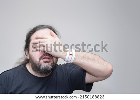 An unshaven man with bracelets in the colors of the Japanese flag pathetically closes his eyes with his palm.