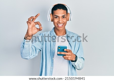 Young african american guy using smartphone wearing headphones doing ok sign with fingers, smiling friendly gesturing excellent symbol 