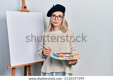 Beautiful caucasian woman drawing with palette on easel stand relaxed with serious expression on face. simple and natural looking at the camera. 