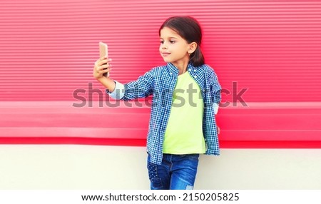 Portrait of happy little girl child taking selfie by smartphone on pink background in the city
