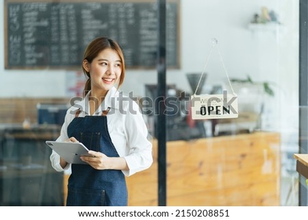 Portrait of happy woman standing at doorway of her store. Cheerful mature waitress waiting for clients at coffee shop. Successful small business owner in casual wearing blue apron standing at entrance Royalty-Free Stock Photo #2150208851