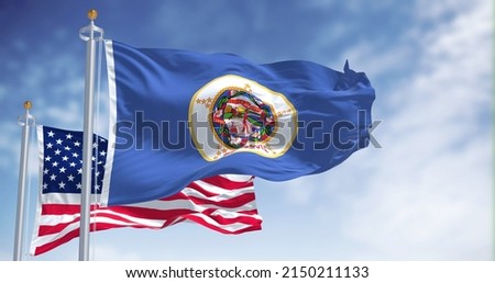 The Minnesota state flag waving along with the national flag of the United States of America. In the background there is a clear sky. Minnesota is a state in the upper Midwestern United States Royalty-Free Stock Photo #2150211133