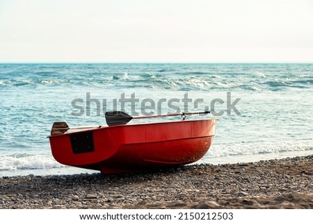 Small red rowboat with sculls lying at shore of the sea Royalty-Free Stock Photo #2150212503