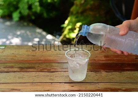 Hand of woman poured water from a clear plastic bottle into a glass filled with ice on a blurred natural background. Humans should drink about 2 liters of water a day to compensate for the water lost 