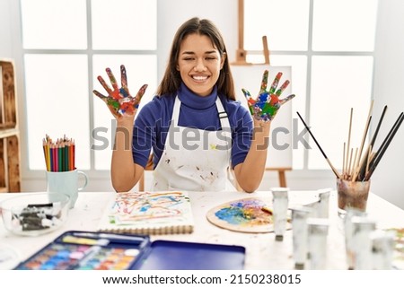 Young brunette woman at art studio with painted hands showing and pointing up with fingers number ten while smiling confident and happy. 