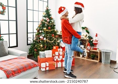Young latin couple hugging and standing by christmas tree at home.