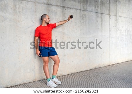Caucasian male sportsman taking a selfie. He is wearing a red T-shirt and blue trousers. Concrete wall background. Spain, Alicante