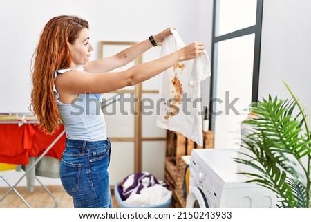 Young redhead woman holding dirty t shirt at laundry room Royalty-Free Stock Photo #2150243933