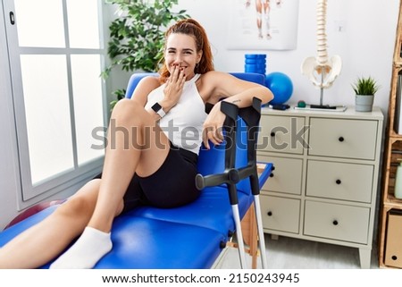 Young redhead woman lying on rehabilitation bed holding crutches laughing and embarrassed giggle covering mouth with hands, gossip and scandal concept  Royalty-Free Stock Photo #2150243945
