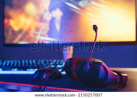 Close up of headset laying on gaming desktop. Selective focus. Monitor and keyboard on background. Professional gaming setup in neon lights ready to use for online esport competition. Cyber sport Royalty-Free Stock Photo #2150257007