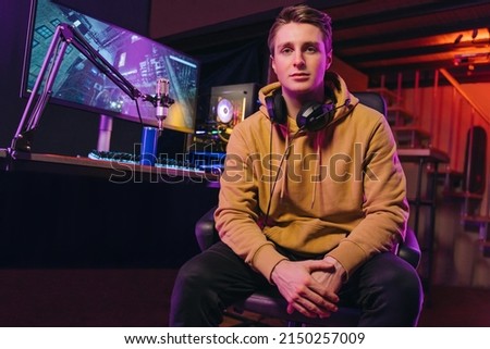 Portrait of young handsome pro gamer streamer wearing headphone smiling at camera while sitting at gaming dest and prepare to live stream. Monitor and microphone with online video game on background