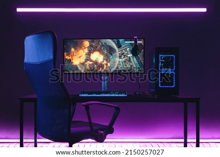 General view of home workplace of pro gamer with professional gaming setup on desktop. Modern powerful PC full RGB light inside, display with shooter game, armchair. Gaming studio of cyber sportsman Royalty-Free Stock Photo #2150257027