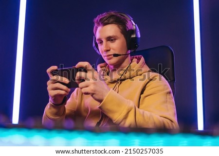 Portrait of caucasian man pro gamer sitting on gaming chair, wearing headset, has live stream while playing mobile game on the smartphone at home. Young cyber sportsman holding cellphone in hands