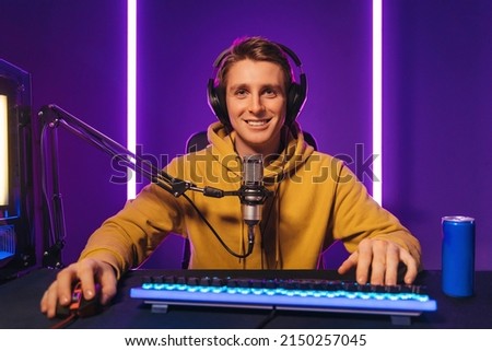 Smiling professional gamer streaming esport tournament, playing online video game at home, using microphone and headphones, drinking energy caffeine drink, enjoying the game, looking at camera  Royalty-Free Stock Photo #2150257045