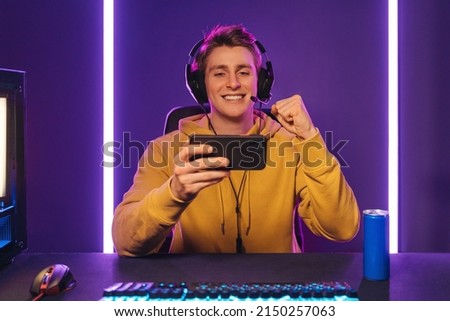 Playing video games on smartphone. Young caucasian handsome man sitting on chair holding cellphone in his hand. Exited streamer wearing headset celebrating victory in online mobile game in neon room Royalty-Free Stock Photo #2150257063