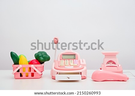 play store, plastic children's toy vegetables in pink basket, cash register, scales on white background, online ordering of products from store