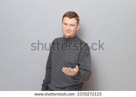Studio portrait of friendly mature man stretching one hand to you, asking your opinion, waiting your decision, offering something, smiling, wearing casual jumper, standing over gray background