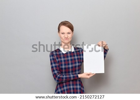 Studio portrait of disgruntled young blond woman holding white blank paper sheet with place for your text and design in hands, wearing checkered dress, standing over gray background