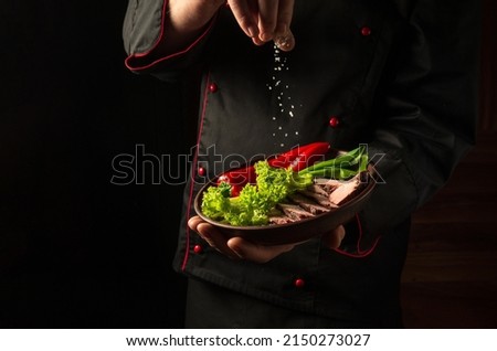 Before serving the order, the chef sprinkles salt on the meat and vegetables on the plate. Food preparation concept on dark background