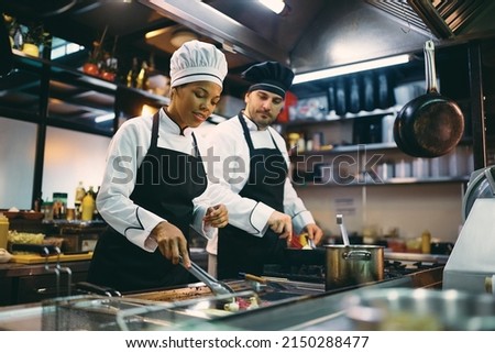 Two professional cooks preparing meal in the kitchen at restaurant. Focus is on African American female chef. Royalty-Free Stock Photo #2150288477