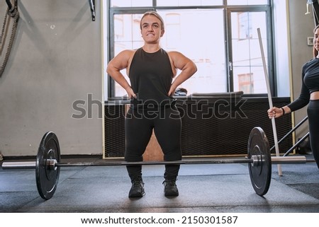 Midget woman standing at the gym with satisfied face while training with barbel near her trainer Royalty-Free Stock Photo #2150301587