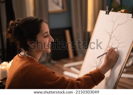 Female with long brown hair pinned up in a bun using pencils sits by candles in the evening. A focused women with the soul of an artist draws a picture of a tree, sketches on a cavas