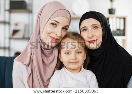 Portrait of happy three generations of muslim women hug and cuddle posing at home together, smiling little preschooler arabian girl young mom and senior grandmother enjoy leisure family weekend Royalty-Free Stock Photo #2150305063