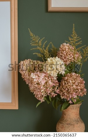 Close up of hydrangea flowers in ceramic vase, wooden picture frame on the wall. Home decoration concept.