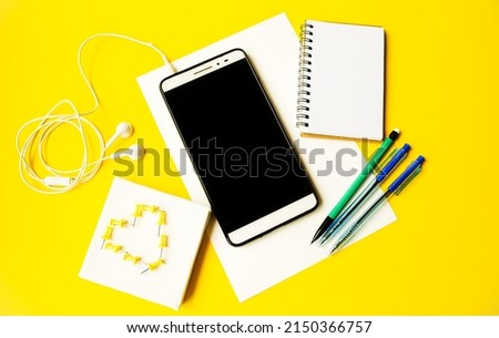 Flat lay on a yellow background lies a mobile phone tablet, a spiral notepad, pens and a pencil, headphones and pins.  Copy space for text, online learning concept, close-up, background image.