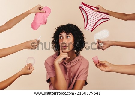 Young woman contemplating the best sanitary choice for her body. Thoughtful young woman looking away while surrounded by hands holding different reusable and non-reusable sanitary products. Royalty-Free Stock Photo #2150379641