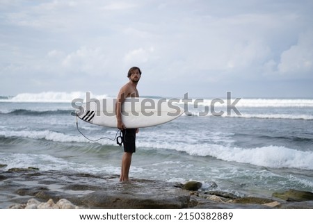 Landscape image of man surfer busy walking on the beach at sunrise, surfboard under his arm with the ocean waves breaking in the background. Young handsome male surfer on the ocean.