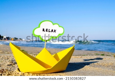 Small origami yellow paper boat with stick and speech bubble with word RELAX stands on sandy beach near sea water and sea waves with white foam close-up. Concept of travel, tourism, vacation, holiday