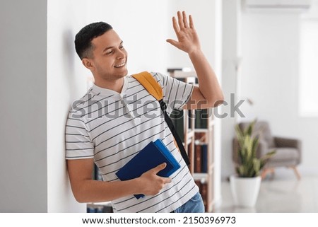 Male student with books waving hand in library