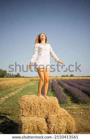 Happy barefoot girl dancing in the agricultural field with haystack and bales after harvesting.
