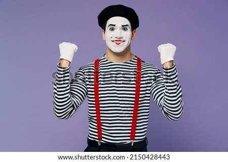 Excited jubilant young mime man with white face mask wears striped shirt beret doing winner gesture celebrate clenching fists say yes isolated on plain pastel light violet background studio portrait