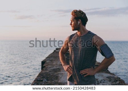 Young strong sporty athletic toned fit sportsman man in sports clothes earphones warm up train with mobile phone akimbo at sunrise sun dawn over sea beach outdoor on pier seaside in summer day morning Royalty-Free Stock Photo #2150428483