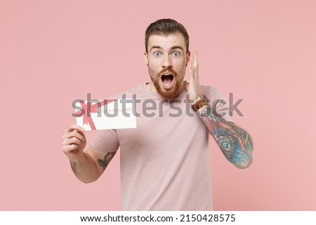 Excited happy young bearded tattooed man wears pastel casual t-shirt hold gift certificate coupon voucher card for store celebrate isolated on pink background. Mock up copy space. Tattoo translate fun