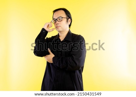 Young asian man wear black t shirt and eyeglasses is suspicious or thinking about something isolated on yellow background 
