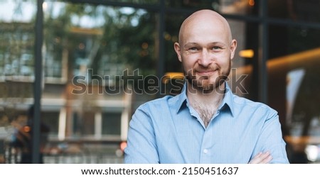 Adult bald smiling attractive man forty years with beard in blue shirt businessman against glass wall of street cafe, banner Royalty-Free Stock Photo #2150451637