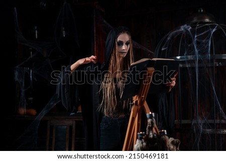 Halloween concept. Witch dressed black hood standing dark dungeon room use magic book for ritual devil calling. Female necromancer wizard gothic interior side view