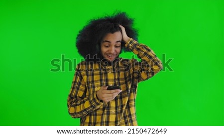 A young man with an African hairstyle on a green background looks at the phone and is happily surprised. Emotions on a colored background