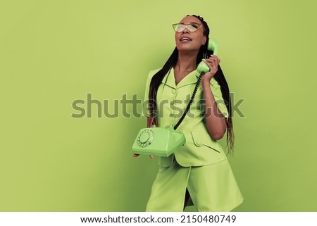 Monochrome portrait of smiling african girl wearing retro style outfit holding vintage phone isolated on green background. Concept of beauty, art, fashion, youth, sales and ads. Copy space for ad. Royalty-Free Stock Photo #2150480749