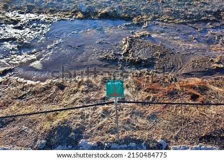 A green sign warning about the dangerous up to 100 degrees high temperature of the water of a small stream flowing in a geothermal vulcanic park near geyser in Iceland, Europe