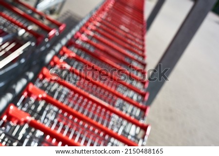 Blurred. A row of shopping carts with red handles from a grocery store (Penny). Gray columns and parking place in the background. View from diagonal above. All out of focus.
