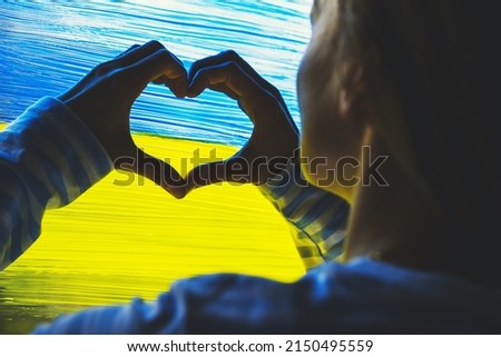 Female hands shaping heart symbol in background of yellow-blue flag of Ukraine on window. Human hands made heart shape on image of flag of Ukraine on glass. Support Ukrainians. Concepts symbol